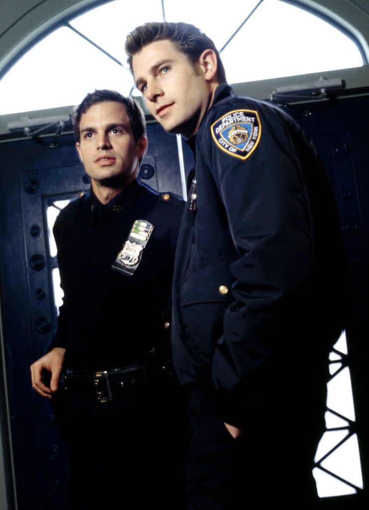 mark ruffalo and derek cecil in character for the beat, both men wear navy blue police uniforms with badges on their chests, they stand inside a set of double doors and look past the camera