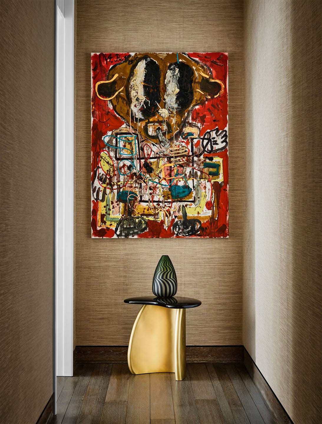 in a hallway with a dark wood floor and light brown textured wallcovering is a small table with a gold base and black top and a glass vase atop it, a piece of abstract art with red background hangs above table