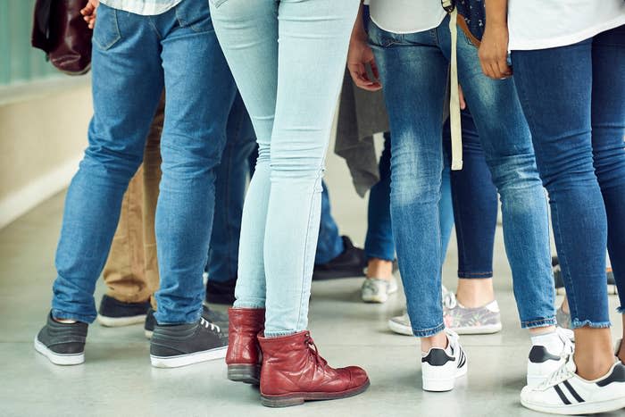 Group of people standing, focusing on their legs and footwear. Jeans and various types of casual shoes can be seen. Names of individuals are not provided