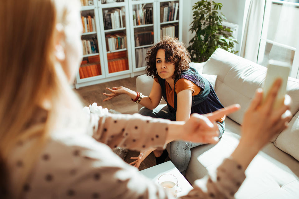 A woman pointing to her phone looking at another woman on the couch