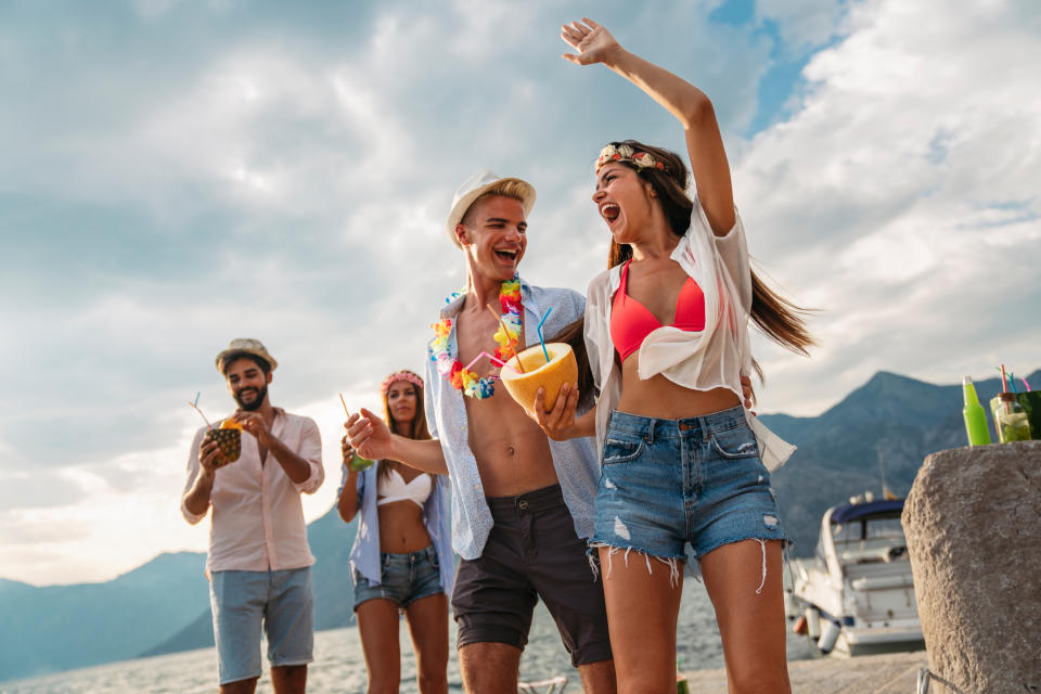 Group of friends enjoying a tropical vacation by the beach, wearing summer clothes, holding drinks, and dancing with a boat and mountains in the background