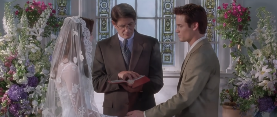 A bride and groom stand at the altar, she has a traditional veil covering her face and he is in a khaki suit - they're surrounded by flowers and an officiant stands between them reading from a book