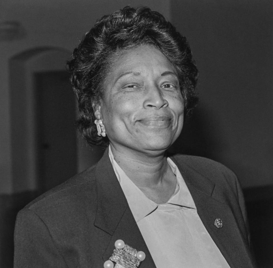 eva m clayton smiles at the camera, she wears a dark colored suit jacket over a lighter colored top along with earrings and lapel pins