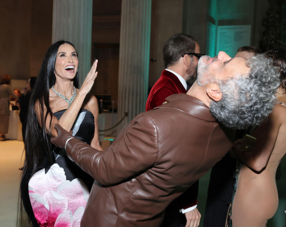 Demian Bichir laughs with an elegantly dressed woman at an event; both engaging warmly in a greeting gesture