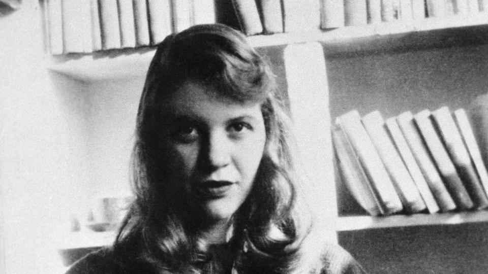 sylvia plath sitting in front of bookshelves and looking ahead for a photograph