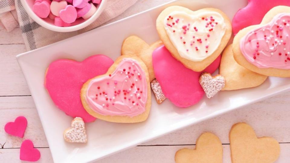 Heart shaped Valentines Day cookies with pink and white icing and sprinkles. Overhead view on a serving plate against a white wood background.