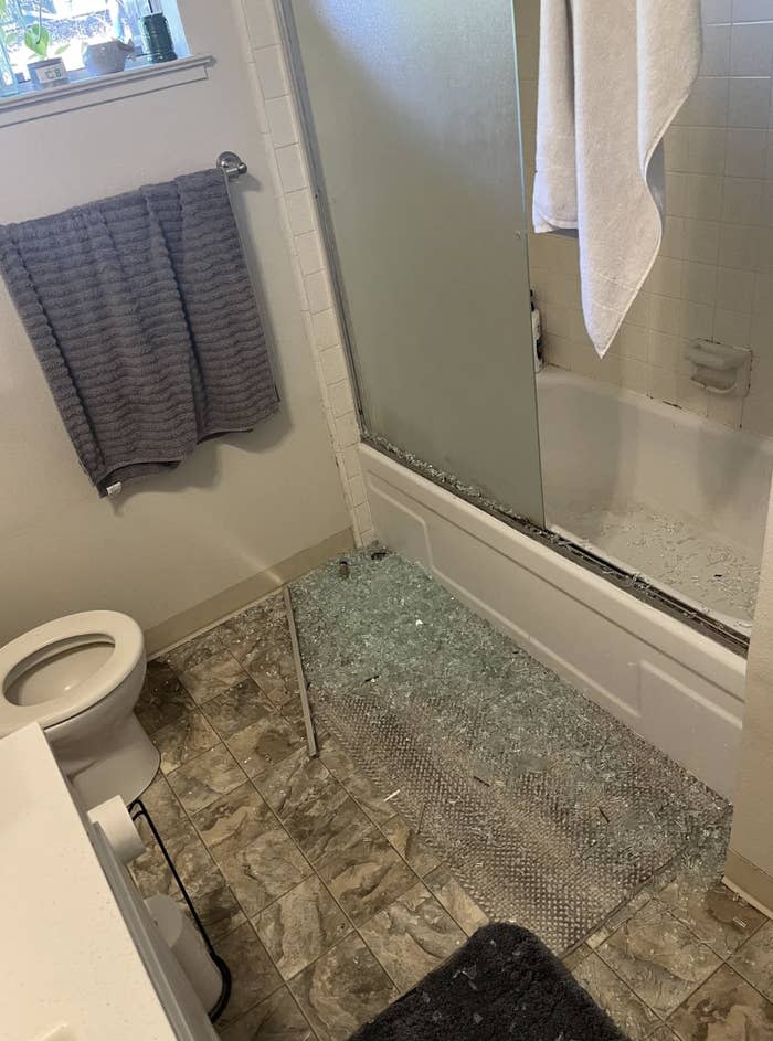 Bathroom with shattered glass door on the floor next to a bathtub; towel hangs on the side