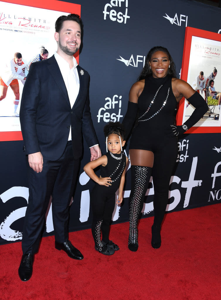 Serena Williams wears a sleeveless dark mini dress with elbow length gloves, Olympia Ohanian wears a dark jumpsuit, and Alexis Ohanian wears a dark suit