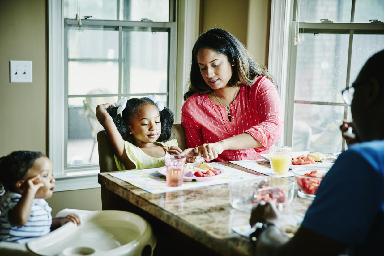 Mother cutting young daughters food while eating breakfast together at dining room table