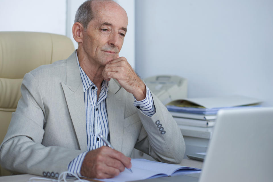 Older man in suit jotting down notes while staring at a laptop screen.