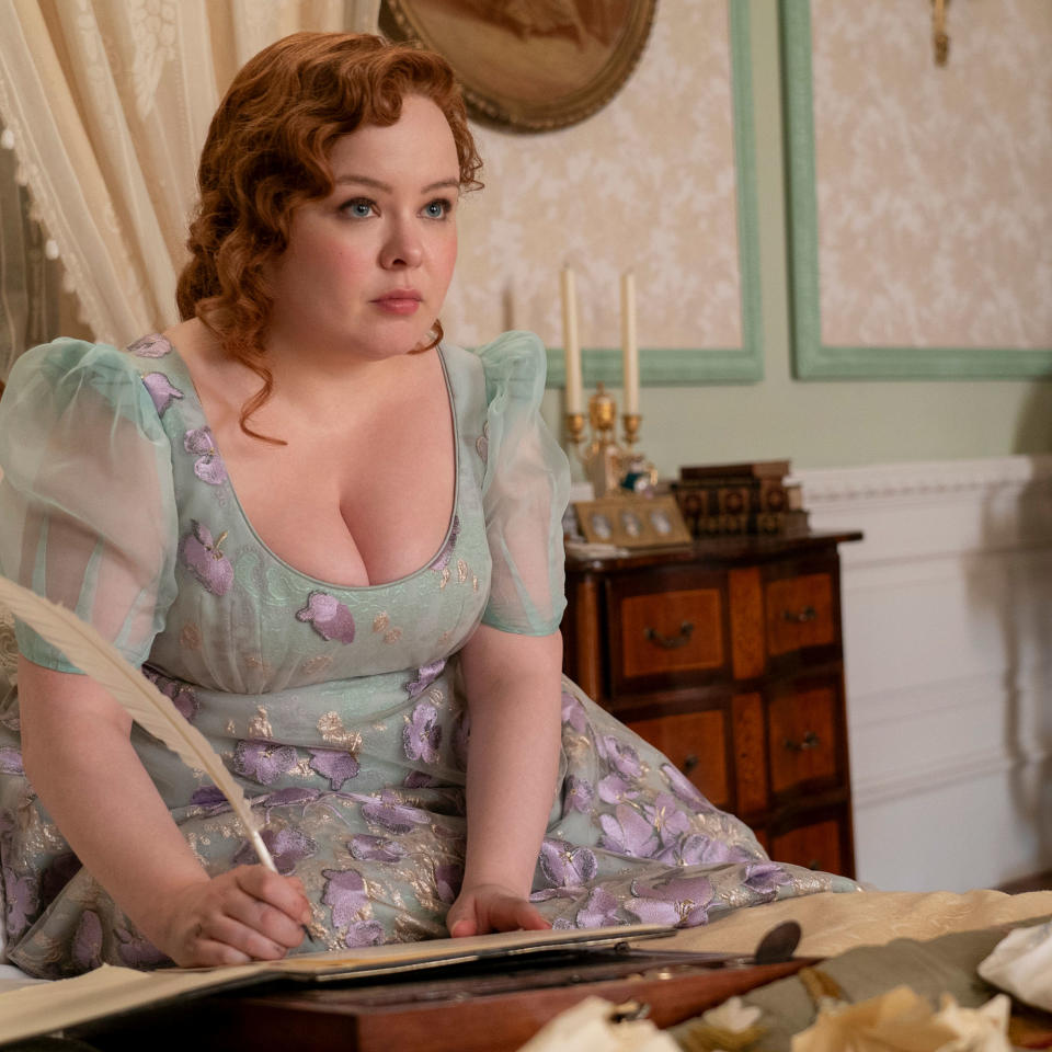 Bridgerton character Penelope sat on bed holding quill over writing desk with drawers and green walls in background
