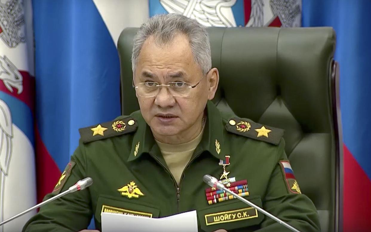 Sergei Shoigu sits before two microphones in front of a flag backdrop.