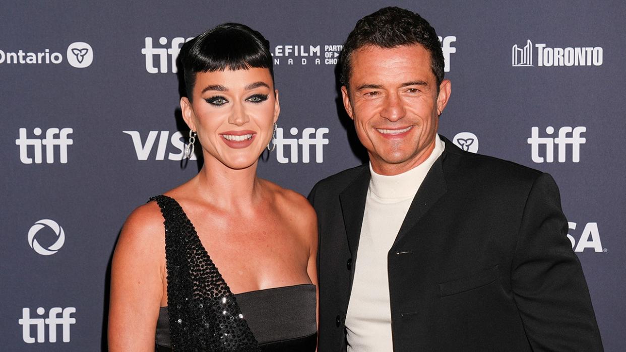 Katy Perry in a one-shoulder sparkly black gown smiles ont he carpet with Orlando Bloom in a white turtleneck and black jacket at the Toronto International Film Festival