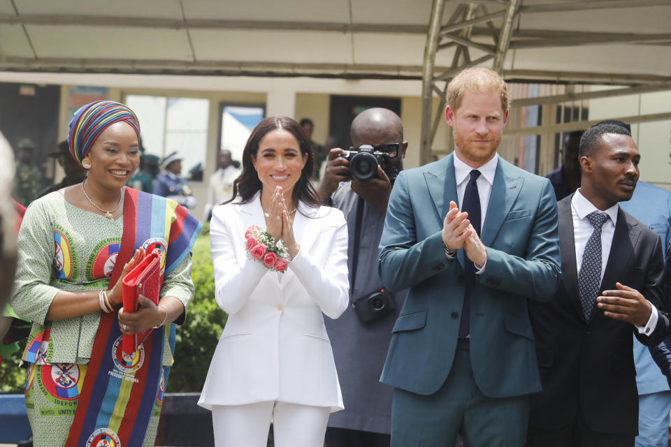 Meghan Markle wears a white suit and a collar necklace standing next to prince harry