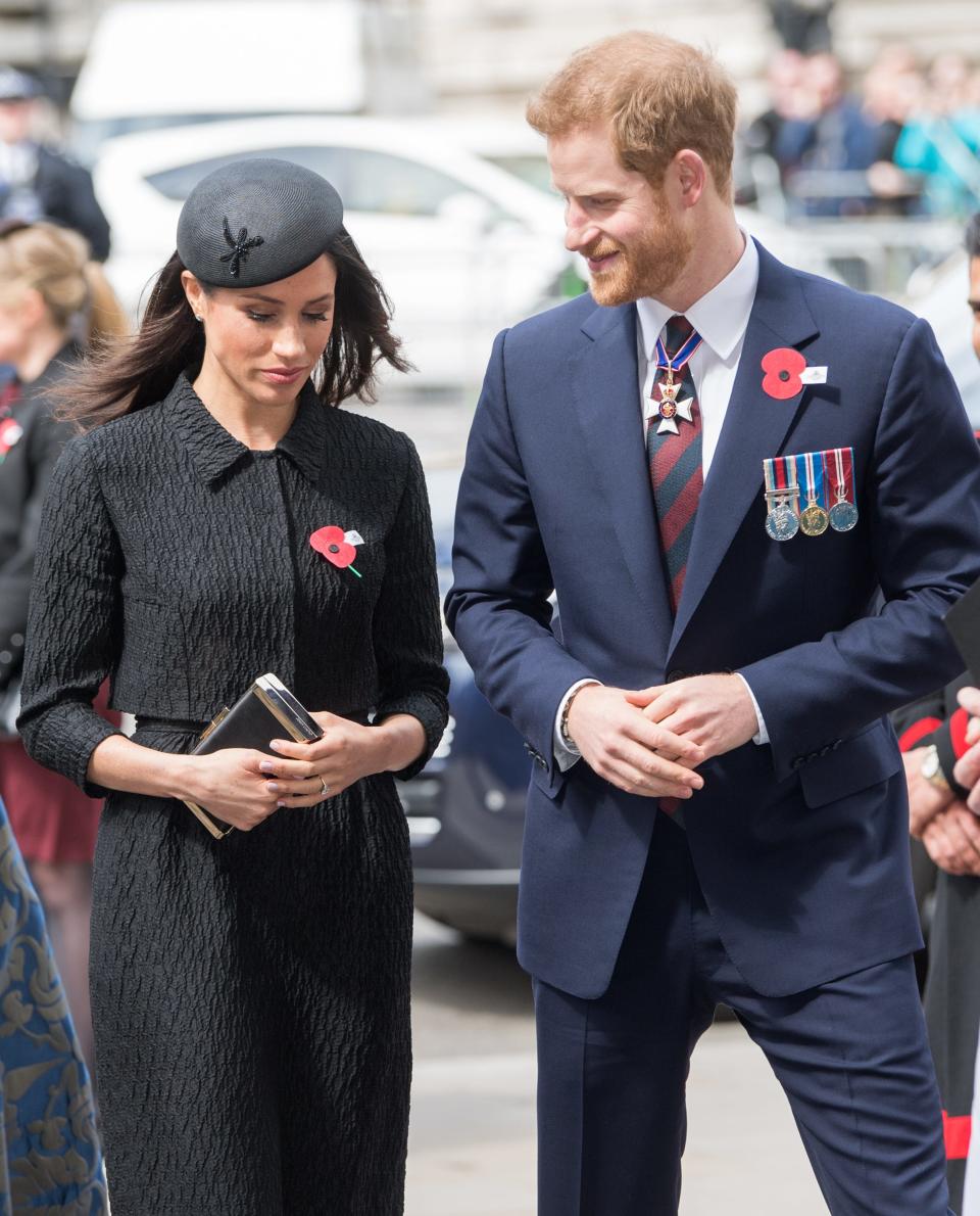 Meghan in a textured black cropped jacket and skirt with a baret and Harry in a navy suit with a navy and red striped tie and both wearing red remembrance poppies.