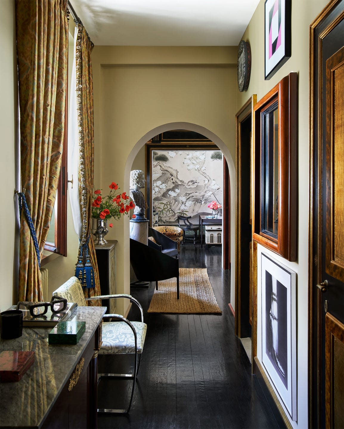 a hallway with dark wood floor, marble topped side table, chair with steel base, curtained window, and multiple framed artworks leads to a rom with twin side chairs, a bust on a plinth, and a japanese style screen