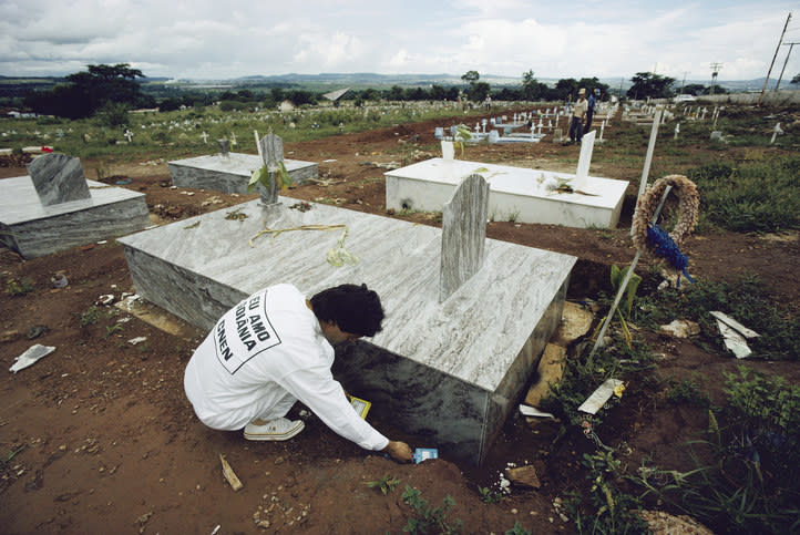 Person in white overalls cleaning a grave marker in a large cemetery. The scene is outdoors with many graves in the background