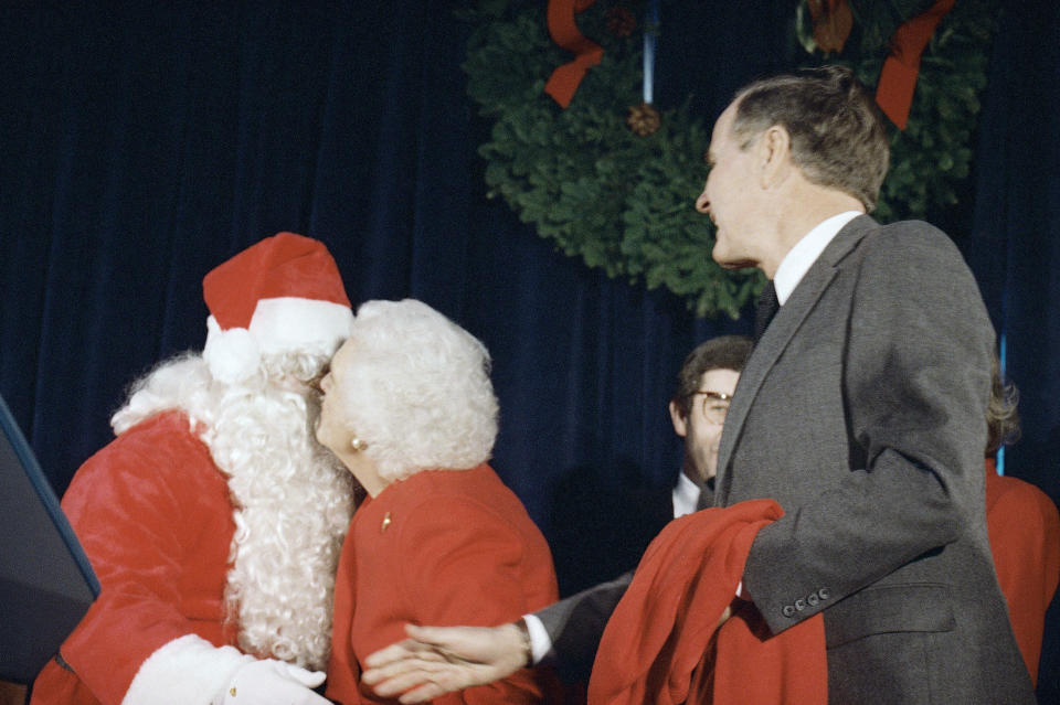 President-elect George H. W. Bush, right, and Mrs. Barbara Bush, kissing Santa, hold sweaters that were given to them at his inaugural headquarters, Monday, Dec. 20, 1988, Washington, D.C. They visited the headquarters to wish all workers happy holidays, and the sweaters were gifts from the staff. (AP Photo/Doug Mills)