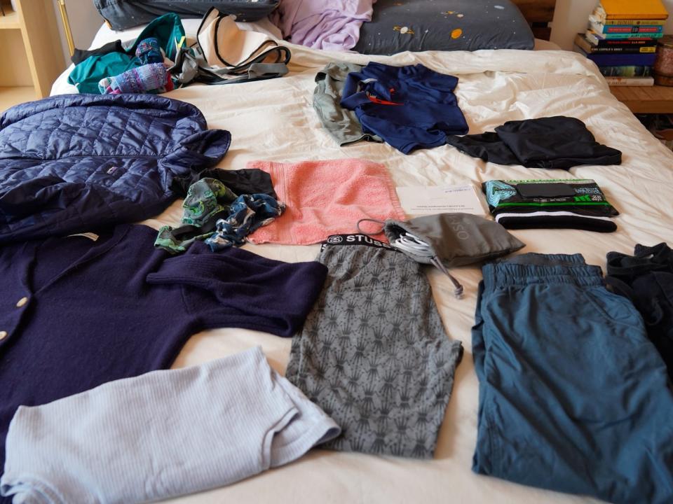 Clothing on a white bed spread including (from left to right): a navy blue jacket and cardigan, a light blue tank top, a pink rag, grey leggings, a short stack of grey and navy blue shirts, a black package of wipes, a black hoodie, and dark teal pants