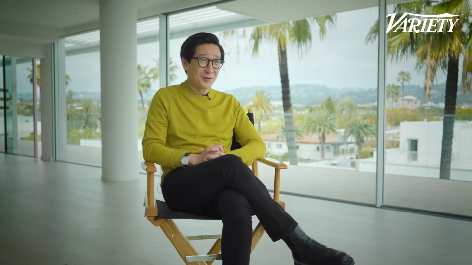 Ke Huy sitting in a director's chair during his Variety interview in a room with floor-to-ceiling windows. There are houses and palm trees outside