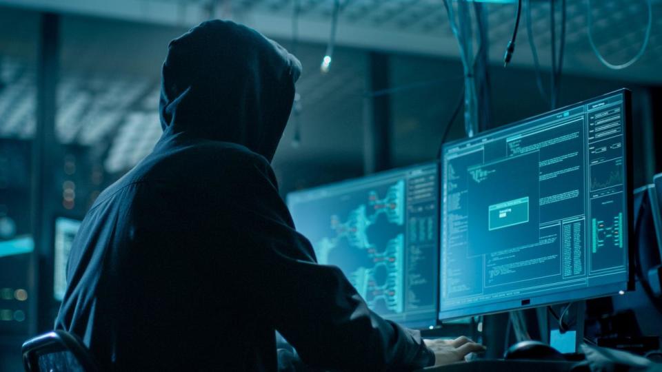 Shot from the Back to Hooded Hacker Breaking into Corporate Data Servers from His Underground Hideout. Place Has Dark Atmosphere, Multiple Displays, Cables Everywhere.