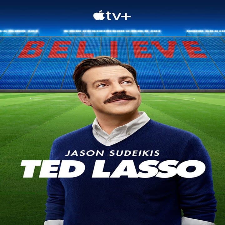 Man with mustache standing in empty soccer stadium, text in stands reads BELIEVE, text across chest reads TED LASSO.