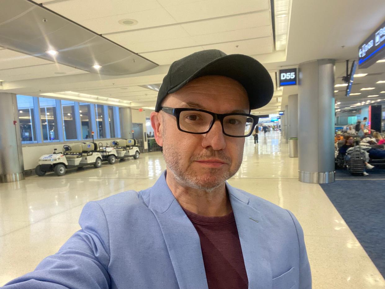 Man wearing black baseball cap, black glasses and blue blazer takes a selfie in an airport