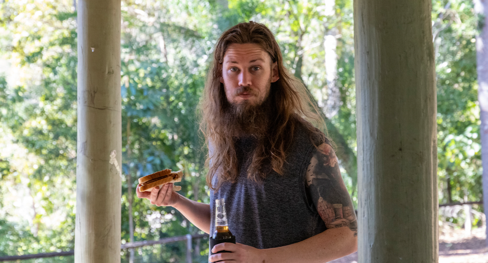 A man with long hair and a beard staring at the camera with a sausage in his hand.