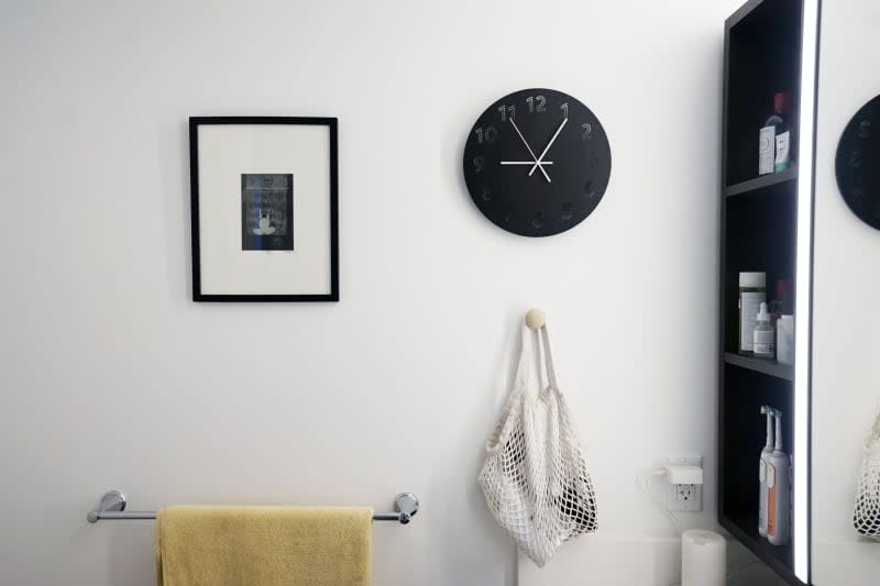 Framed artwork next to a modern wall clock in a white-walled bathroom.