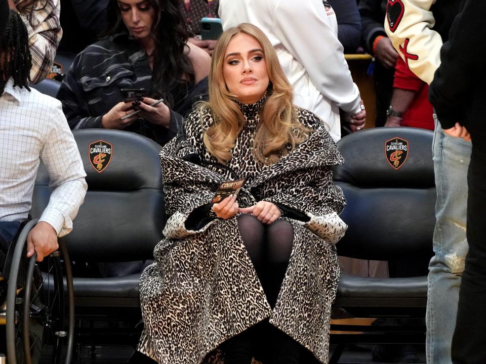 Adele with an annoyed or bored face sitting alone in the front row of a basketball court.