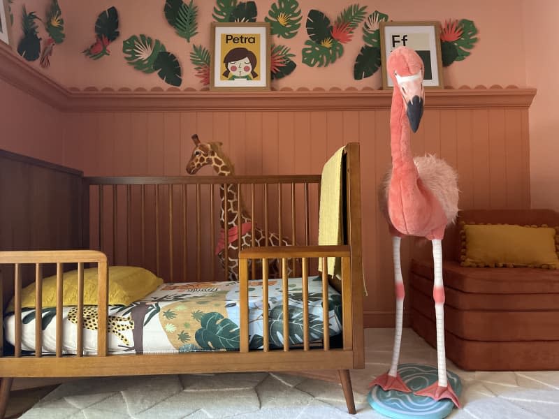 large human life size flamingo statue wearing sunglasses, beige patterned rug, 3/4 wall paneling, wall trim, pink and dusty rose walls, dusty rose velvet arm chair, mid century inspired wood crib, small child's bed, picture shelf, leafy mural on wall, big giraffe stuffy