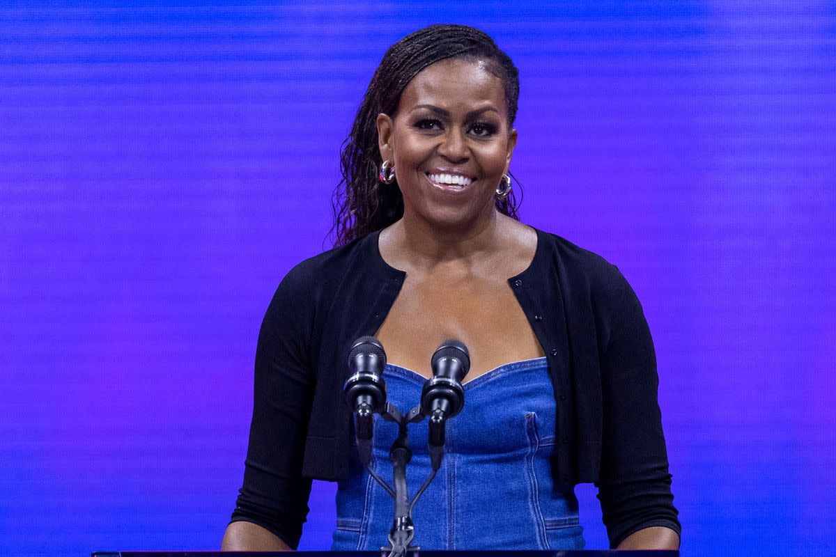 A Black woman wearing a black cardigan over a top made out of jean material smiles at a podium. 