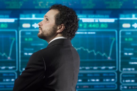 An investor in a suit, looking at various digital charts on a large ticker board.