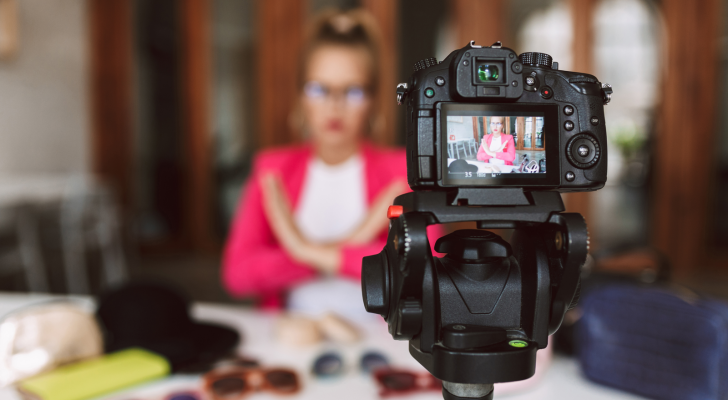 A woman in a pink jacket sits at a table inside while filming herself on camera