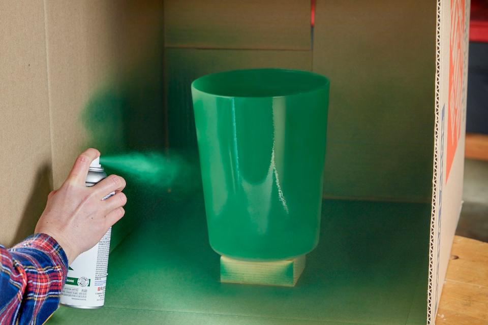 Using green spray paint on plastic bin in cardboard box.