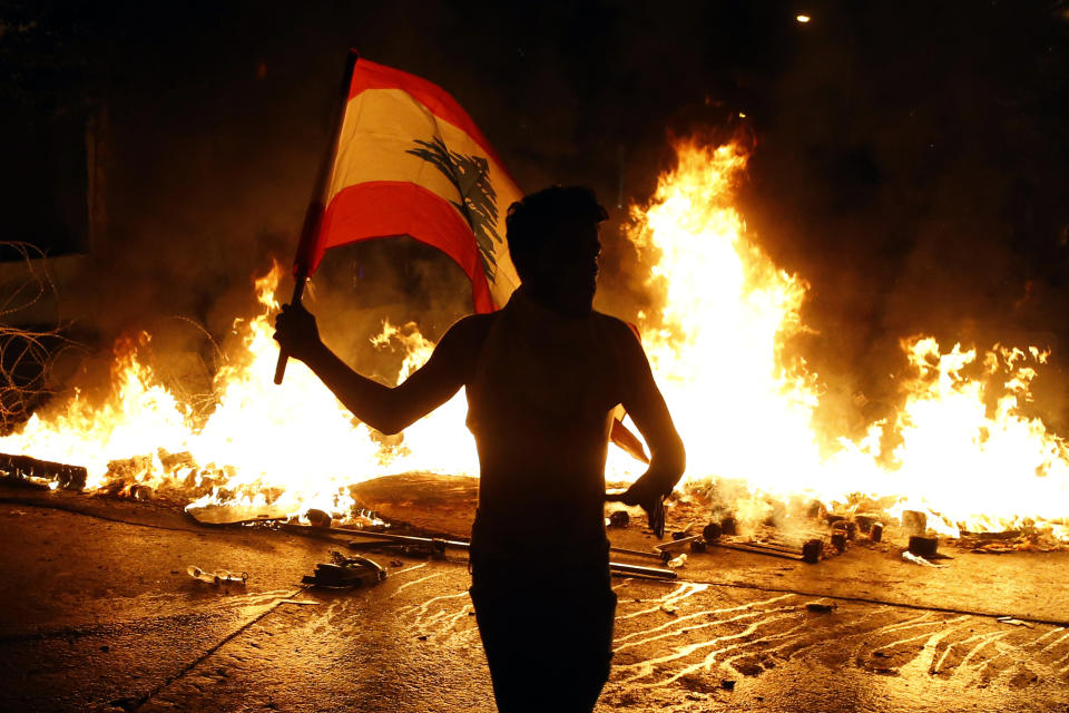 An anti-government protester waves a Lebanese flag in front of a barricade on fire on a road leading to the parliament building, during ongoing protests against the government, in Beirut, Lebanon, Wednesday, Nov. 13, 2019. Lebanese protesters said they will remain in the streets despite the president's appeal for them to go home. (AP Photo/Bilal Hussein)