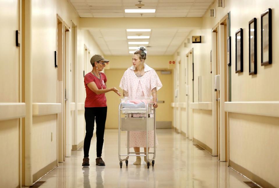 Two women, one pushing a newborn baby in a cart, walking in a hospital hallway.