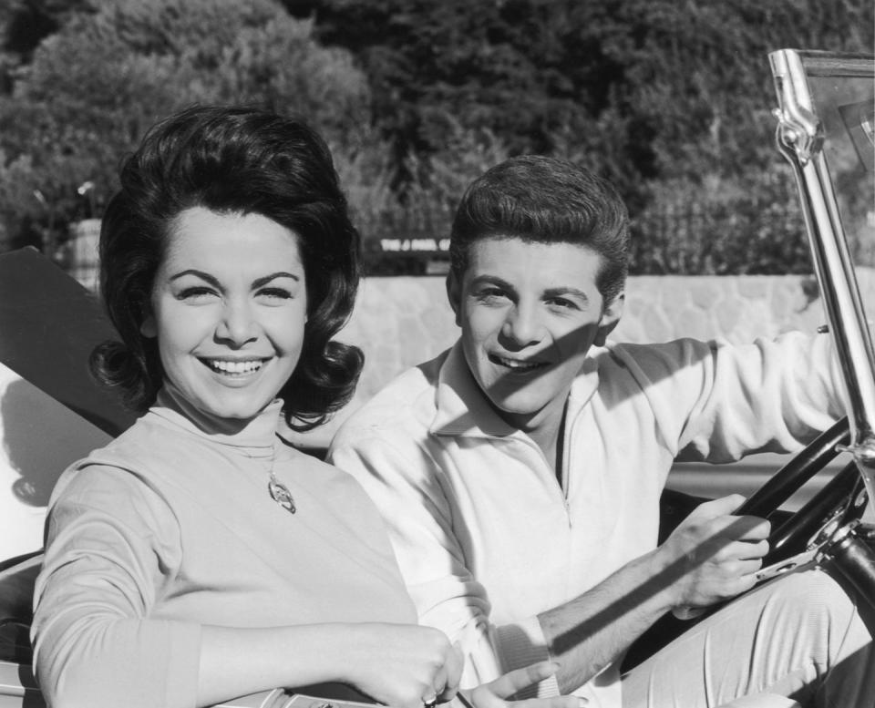 annette funicello and frankie avalon smile while sitting in the front seat of a convertible car, she wears a turtleneck long sleeve shirt with a necklace, he wears a quarter zip sweater