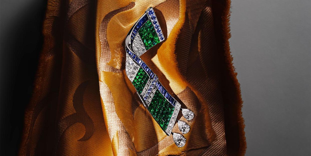 flat emerald and diamond bracelet with blue sapphire edging and three teardrop diamonds on one end displayed on a golden satin patterned cloth