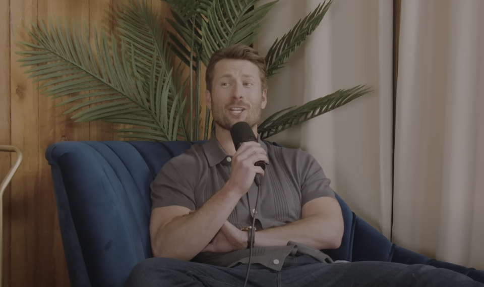 Man sitting in a blue chair, holding a microphone, speaking. There's a green plant behind him