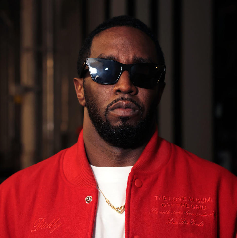 Sean Combs, wearing sunglasses and a red jacket, attends an event. The jacket features the embroidered text, "The Love Album: Off the Grid."