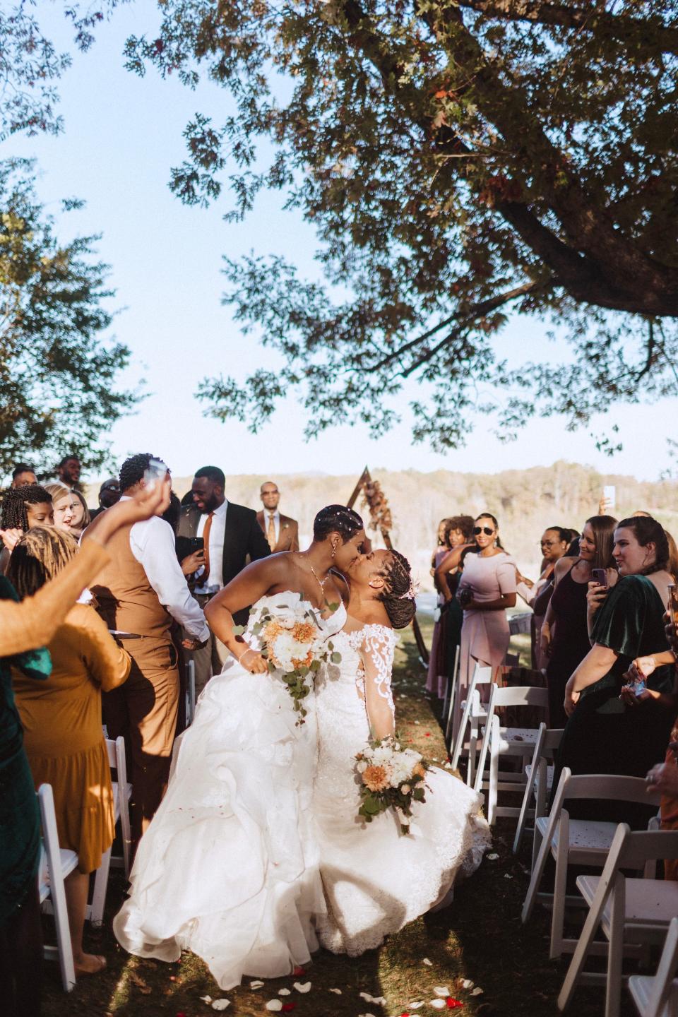 Two brides kiss as they exit their outdoor wedding ceremony.Their friends stand and celebrate them.