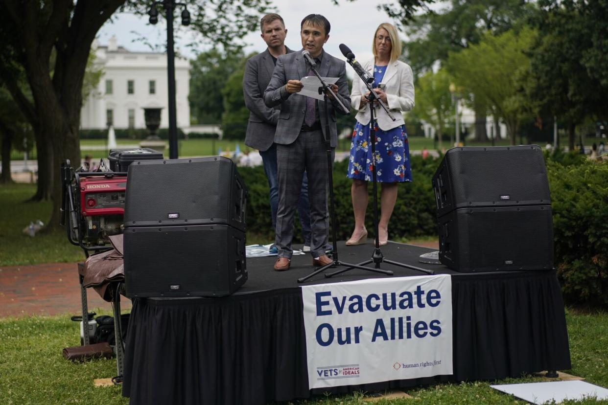 A man speaks while standing on a platform bearing a sign that says, "Evacuate Our Allies," with a man and a woman behind him