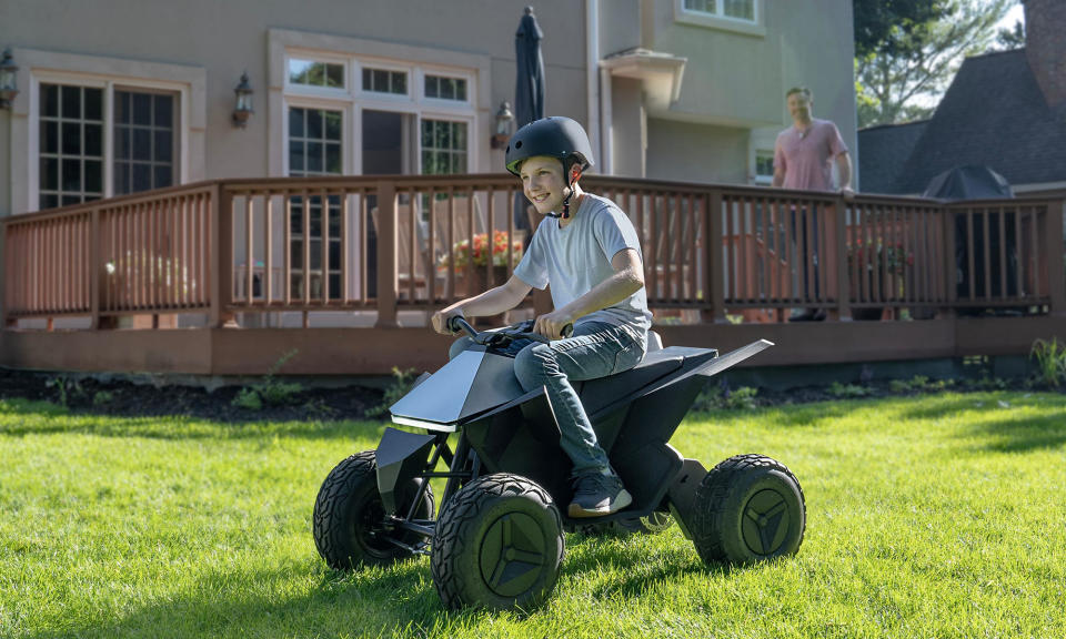 Lifestyle marketing photo of a boy gleefully riding a Tesla Cyberquad for Kids ATV. His father beams from behind as he stands on the family's sunny suburban porch.