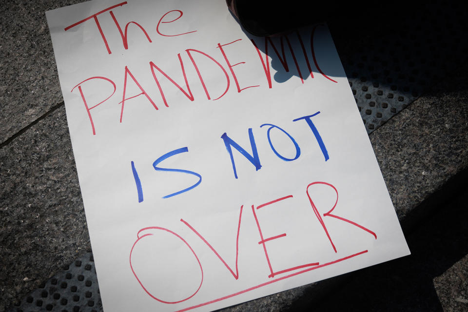 A protest sign on the ground reads "THE PANDEMIC IS NOT OVER" in red and blue ink