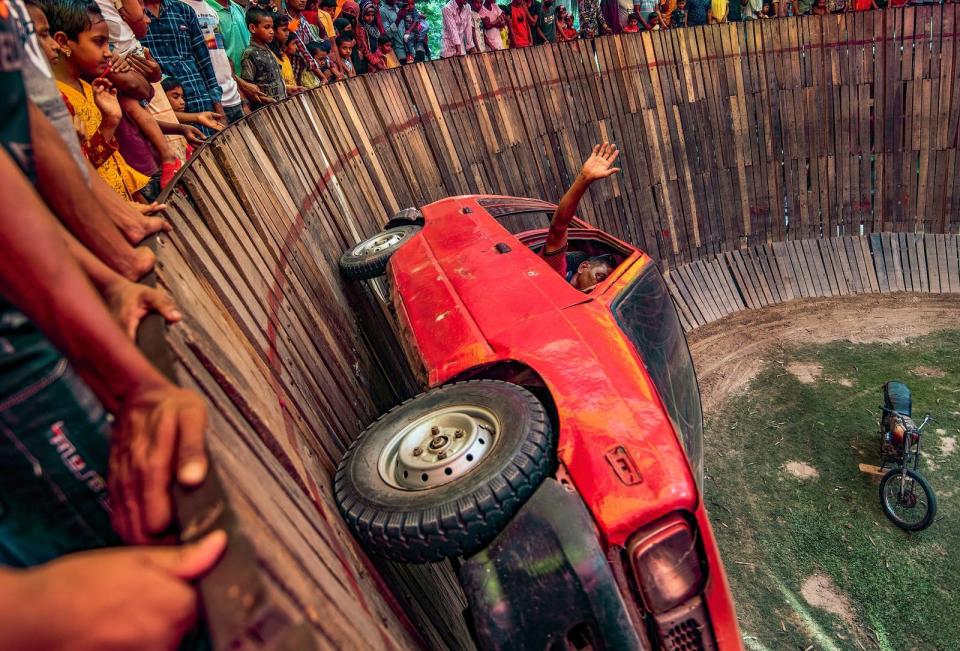 A man waves at the crowd from a red car that is driving around the "Well Of Death," vertically to the ground.