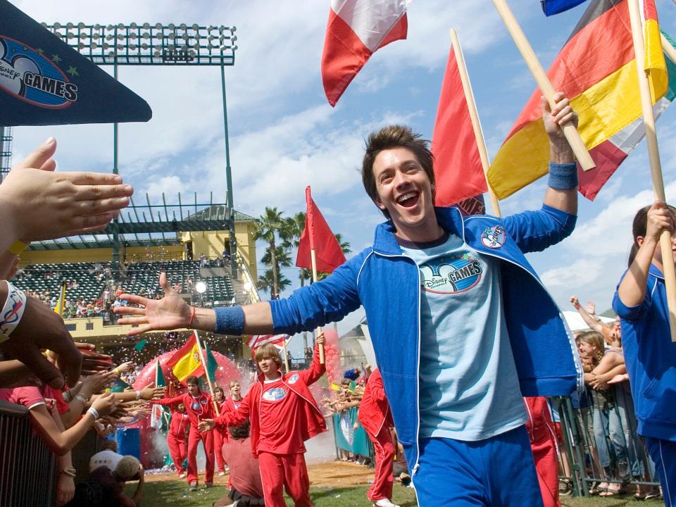 Roger Gonzalez-Garca wearing blue sweatsuit with hand out to high-five fans and flag raised in other hand at disney channel games in 2007