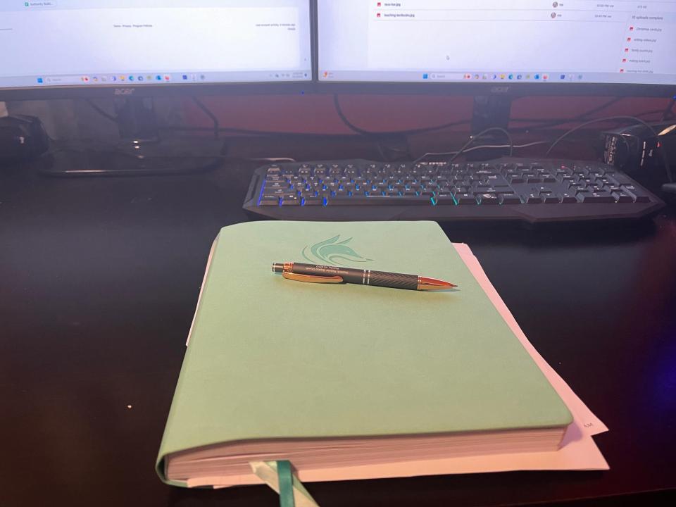 green journal with a pen resting on top sitting on a desk in front of two computer monitors