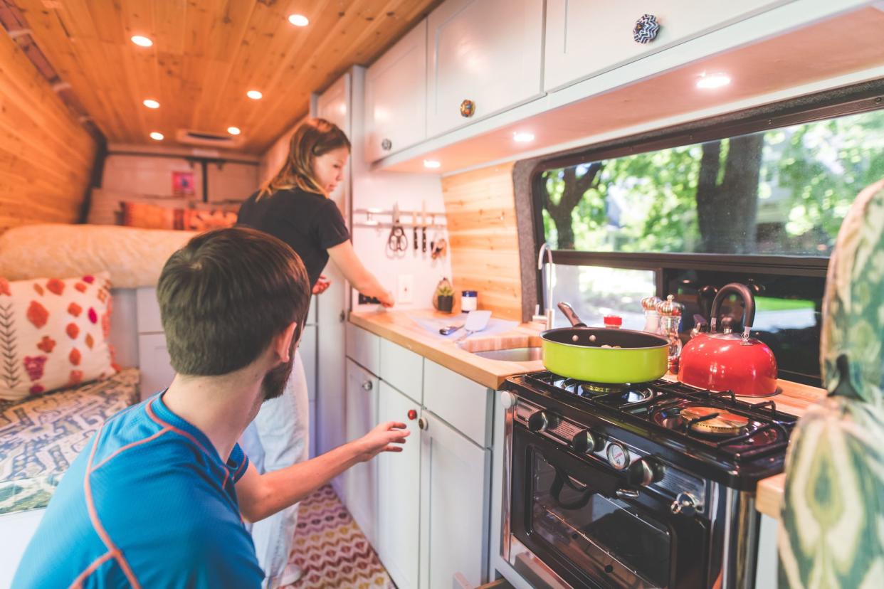 A young couple live in a van. They're making breakfast and she is standing by the sink while he reaches to pick something up off the counter.