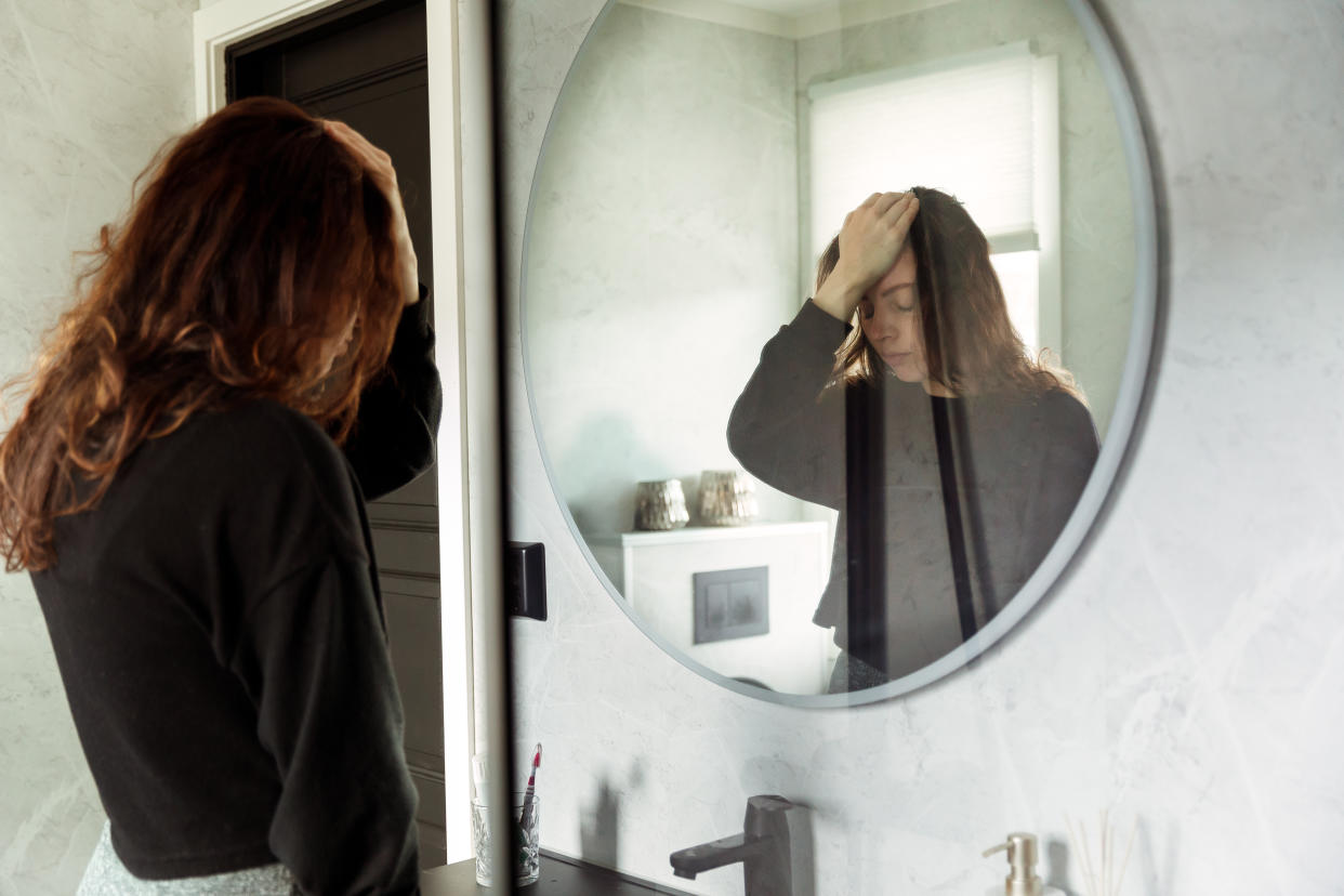 Depressed Woman holding head and looking down in front of bathroom Mirror. The long-term effects of alcohol consumption on the body can vary, and some changes may be reversible to some extent if alcohol use is discontinued and a healthier lifestyle is adopted. (Image via Getty)